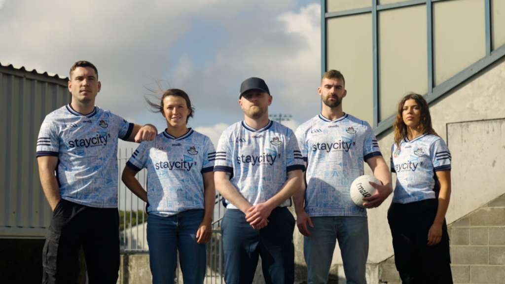Five people standing outdoors wearing light blue Dublin GAA jerseys, with one person holding a football beside a large building.
